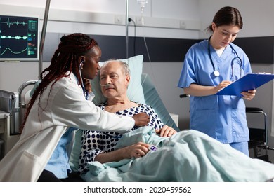 African Doctor Specialist Using Stethoscope Listening Senior Man Heart, Laying In Bed Breathing With Help From Oxygen Tube, And Medical Nurse Is Taking Notes On Clipboard.