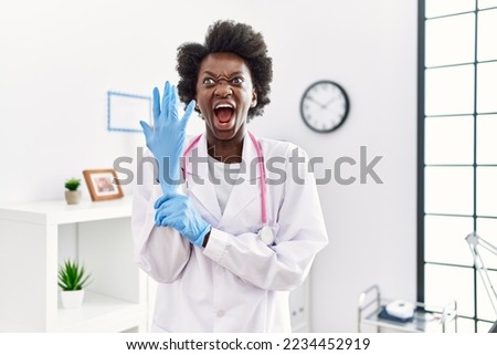 African doctor woman wearing latex gloves at medical clinic angry and mad screaming frustrated and furious, shouting with anger. rage and aggressive concept. 