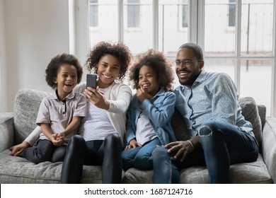 African Ethnicity Couple With Little Kids Resting On Couch Having Fun Holding Smart Phone Using New Free Cool Application, Making Self-portrait Capture Moment, Enjoy Distant Talk By Video Call Concept