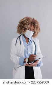 African Ethnicity Doctor With Curly Hair In A Medical Mask In A Lab Coat, Writes Information To The History Of The Disease On A Clipboard Isolated On Grey Background . Concept: Medicine, First Aid