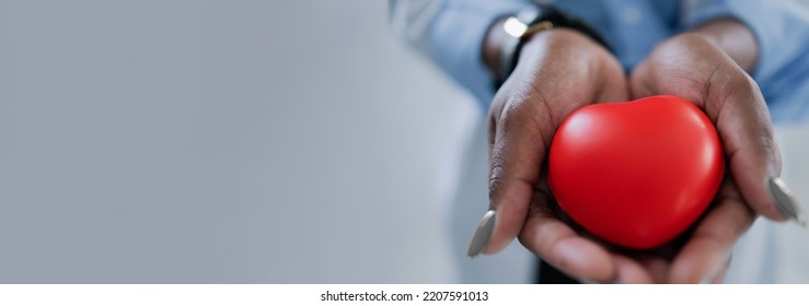 African Ethnicity Doctor Hands Hold A Red Heart On A Gray Background, Baner. Concept: Cardiology, Donation, Mindfulness, Wellbeing, Family Insurance And CSR Concept, World Heart Day, World Health Day