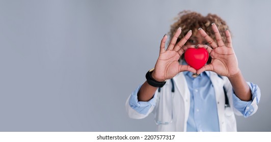 African Ethnicity Doctor Woman With Curly Hair In A Lab Coat Holding A Red Heart On A Gray Background Mocap Place For Design, Baner. Concept: Medicine, Doctor, Ambulance, Cardiology, Donation