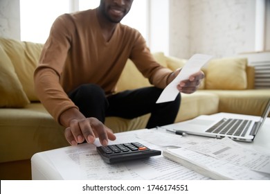 African ethnicity man sitting on couch calculates digits using calculator planning family budget check bills cheques, heap of bill financial papers documents and notebook on coffee table close up view - Powered by Shutterstock