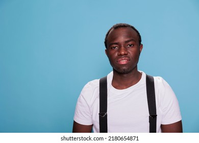 African Ethnicity Man Standing On Studio Blue Background While Looking At Camera. Cool Looking Young Adult Person Wearing White Tshirt And Suspenders Posing Alone Without Face Expression.