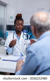 African Ethnicity Woman With Doctor Job Looking At X Ray Scan In Hand While Discussing With Senior Ill Patient At Healthcare Clinic. Black Medic Holding Radiography For Old Man With Disease