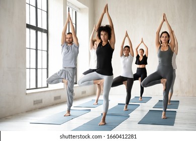 African ethnicity woman yoga coach performs asana standing in Tree Pose or Vrikshasana balancing position with group of multiracial millennial people during morning session, exercise improve stability - Powered by Shutterstock