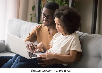 African Family Dad And Child Daughter Using Laptop Computer At Home Sitting On Sofa, Black Father Helps Kid Girl With Homework Having Fun Looking At Pc Screen Doing Online Shopping, Study In Internet