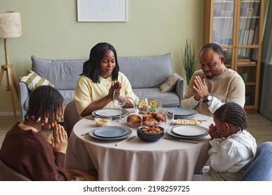 African Family Of Four Sitting At Dining Table And Praying Before Holiday Dinner In Living Room