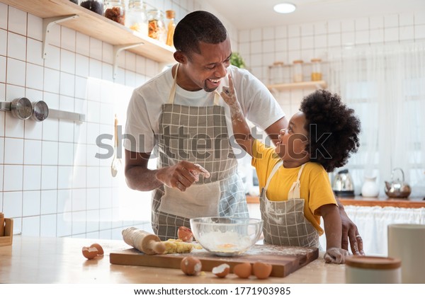 African family have fun cooking baking cake or
cookie in the kitchen together, Happy smiling Black son enjoy
playing and touching his father nose with finger and flour while
doing bakery at home.