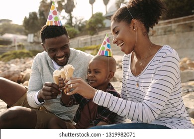 African Family With Party Hats Eating Ice Cream On The Beach. Man And Woman With Son Enjoying Eating Ice-cream. Family Enjoying Son's Birthday With Ice Cream Outdoors.