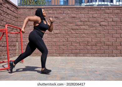 African Fat Woman Jogging During Cardio Workout For Burning Calories Outdoors In City At Day Time, Side View, Copy Space. Young Obese Lady In Black Sportive Tracksuit Is Running Forward, Focused.