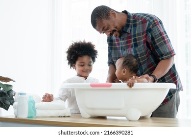 African Father With Beard Bathing Adorable Newborn Baby Daughter In Bathtub With Sponge At Home. Child Boy Help Dad Cleaning His Little Sister In Bath. Kid Hygiene And Cleanliness Care With Family