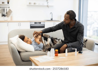 African father cares for sick son by checking temperature with thermometer at home. Scene depicts parent-child bond, concern, and nurturing in light-filled living room. - Powered by Shutterstock