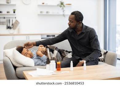 African father checks temperature of ill son resting on couch. Child holds teddy bear, wrapped in blanket. Family scene depicts care and concern in modern home environment. - Powered by Shutterstock