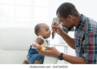 African Father Feeding Adorable Toddler Baby Girl With Spoon While Sitting On Sofa At Home And Kid's Face Mess Up With Food. Little Child Care And Relationship Of Dad And Little Daughter Concept