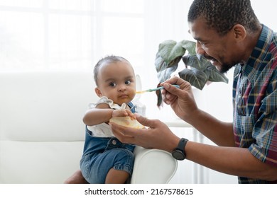 African Father Feeding Adorable Toddler Baby Girl With Spoon While Sitting On Sofa At Home And Kid's Face Mess Up With Food. Little Child Care And Relationship Of Dad And Little Daughter Concept