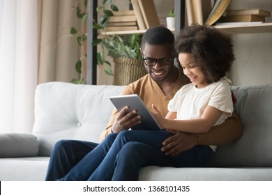 African father holds on lap daughter sitting resting on sofa in living room using digital tablet holding mobile computer watch cartoons educational online program, buying shopping via internet concept