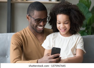 African father and little mixed race daughter sitting on sofa spend time together dad holds smartphone showing to kid new app game, make call take selfie. Having fun at home, modern technology concept