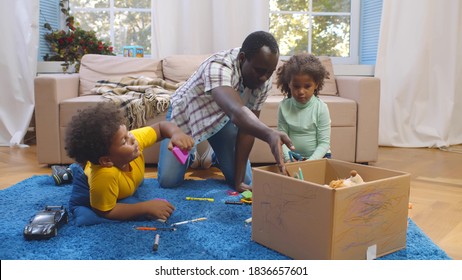 African father and two preschool children sitting on carpet and putting toys in box. Portrait of black dad helping toddler son and daughter cleaning up after playing in living room