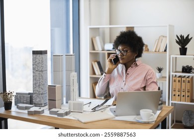 African female architect in office setting communicating on smartphone about city architectural project. Professional environment with model buildings, laptop, and documents. - Powered by Shutterstock