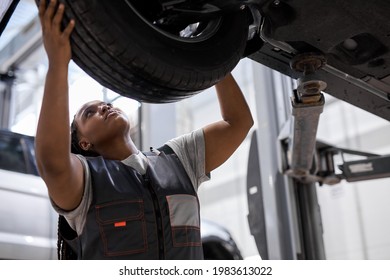 African female auto mechanic changing wheel tire in car in garage, side view. Beautiful black lady in overalls is concentrated on work, carefully adjusting repairing - Powered by Shutterstock