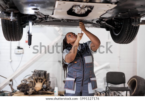 African female auto mechanic fixing car, mechanic\
repairing car on lift in garage. attractive confident black afro\
american lady in overalls concentrated on work, repair using\
instruments tools