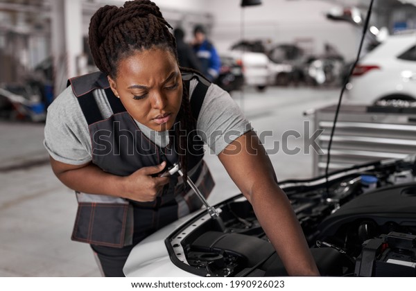 African Female Checks an\
Engine Breakdown. Car Service Employee Woman Fix the Engine\
Component. Modern Clean Workshop. Black Woman In Overalls Looks\
Concentrated