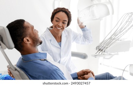 African female dentist turning on lamp before treatment, smiling to male patient - Powered by Shutterstock