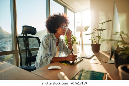 African female designer busy working at her desk. Creative woman working with graphics tablet and looking at desktop monitor in office.