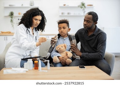 African female doctor checks temperature of young boy at home. Boy holds teddy bear and sits next to African male adult who looks concerned. Scene conveys care, family support, and healthcare. - Powered by Shutterstock