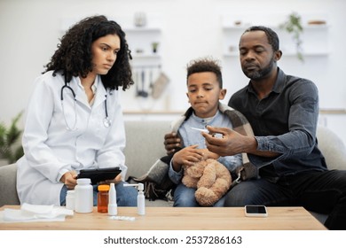 African female doctor consulting African male adult and young boy at home. Doctor holds tablet, father checks thermometer, boy holds teddy bear. - Powered by Shutterstock