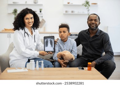 African female doctor displaying lung x-ray on tablet to little sick boy with teddy bear and African father at home. Background shows modern home interior with healthcare items on table. - Powered by Shutterstock