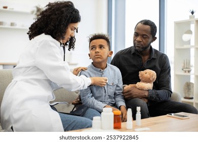 African female doctor examines young boy's heartbeat and lungs at home. Father attentively observes while holding child's teddy bear, forming a comforting scene. - Powered by Shutterstock