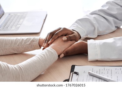 African female doctor holding hand supporting caucasian woman patient. Kind ethnic professional physician give empathy concept encourage reassure infertile patient at medical visit, close up view.