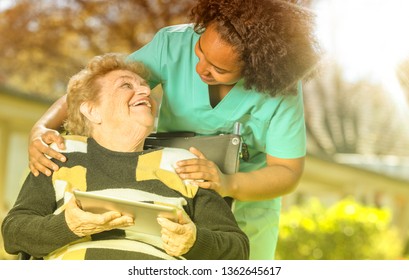 African female doctor playing and smiling with mature elderly woman on wheelchair in the garden using tablet. Retirement community concept