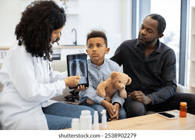 African female doctor showing x-ray of lungs on tablet to little sick boy sitting with father at home. Young boy looks concerned while holding a teddy bear. - Powered by Shutterstock