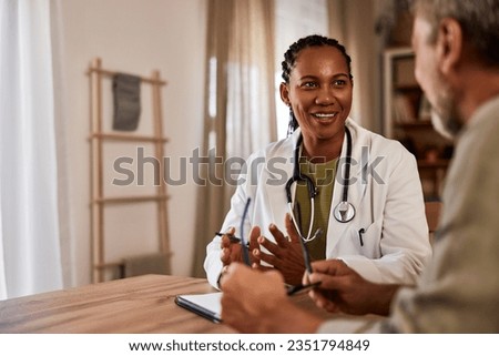 An African female doctor sitting at the wooden table with notes and talking with a patient during a home visit.