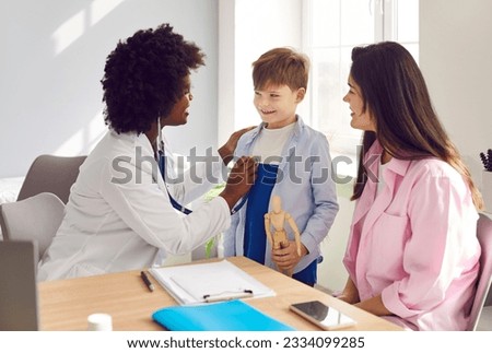 African female pediatrician examining boy patient with stethoscope. Cute boy visiting doctor with his mother. Black woman general practitioner checking heart and lungs of kid at pediatric checkup