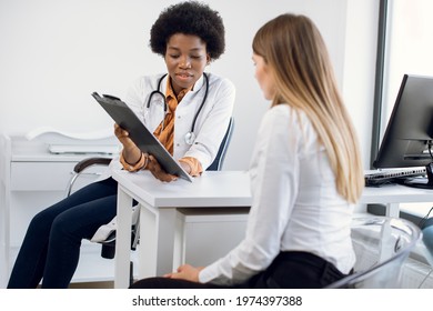 African Female Physician And Young Girl Patient Discussing Something While Sitting At The Table. Medicine And Health Care Concept. Doctor Shows Clipboard With Information For Patient