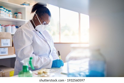 Afrikanische Technikerin in Schutzmantel und Gesichtsmaske, die an einem Tisch im Labor steht und Daten über Proben schreibt