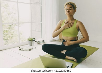 African female yoga teacher having online lesson sitting on green mat in light room showing pranayama techniques, hands on her chest and belly, looking concentrated and focused on body feelings