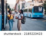 African female young friends waiting at a bus stop and talking cheerfully. Black woman tourists in new town. Best friends traveling together in a city. African american woman using public transport.