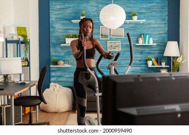 African Fit Strong Woman Doing Cardio Exercise On Eliptical Machine, In Home Living Room Looking At Tv, Watching Instructions Holding Remote Control. Exercising Dressed In Sportwear.