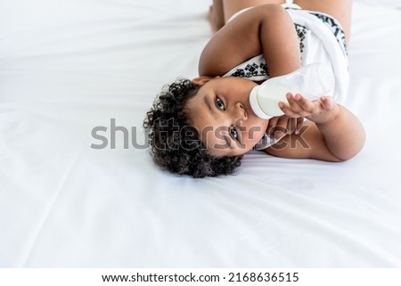 African girl age 2 year and 6 months, lying on a white bed and sucking milk from plastic bottles, with white background, to Child health and food concept.