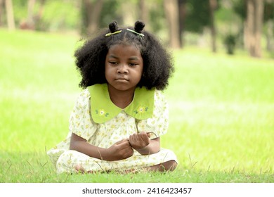 African girl with black curly hair expressing boredom face while setting on green grass meadow garden. Bored child serious thinking during spending time outdoors in summer park. Tired kid sit and rest - Powered by Shutterstock