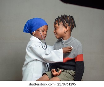 African Girl Child With Nose Mask And Stethoscope, Playing Medical Expert Like Doctor And Nurse With Kid Patient Sitting In Front Of Her