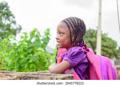 An African Girl Child In School Uniform And With A Backpack, Resting Her Hands On A Low Fence