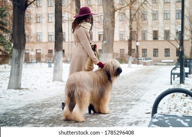An African girl in a coat and hat holds a briard dog on a leash, standing on a winter city street.