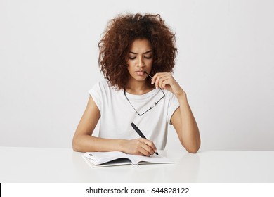 African Girl Thinking Writing In Notebook Sitting At Table Over White Background. Copy Space.