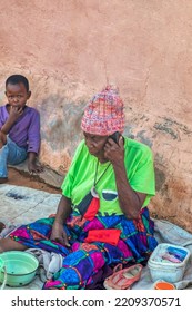 African Granny Talking On The Phone In The Yard, The Grandson Next To Her